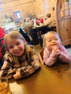 Two sisters sit at a table in a restaurant. The youngest girl has Down syndrome