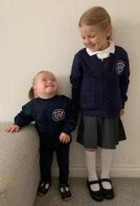 Two sisters stand against a wall in their school uniform. The youngest child has Down syndrome