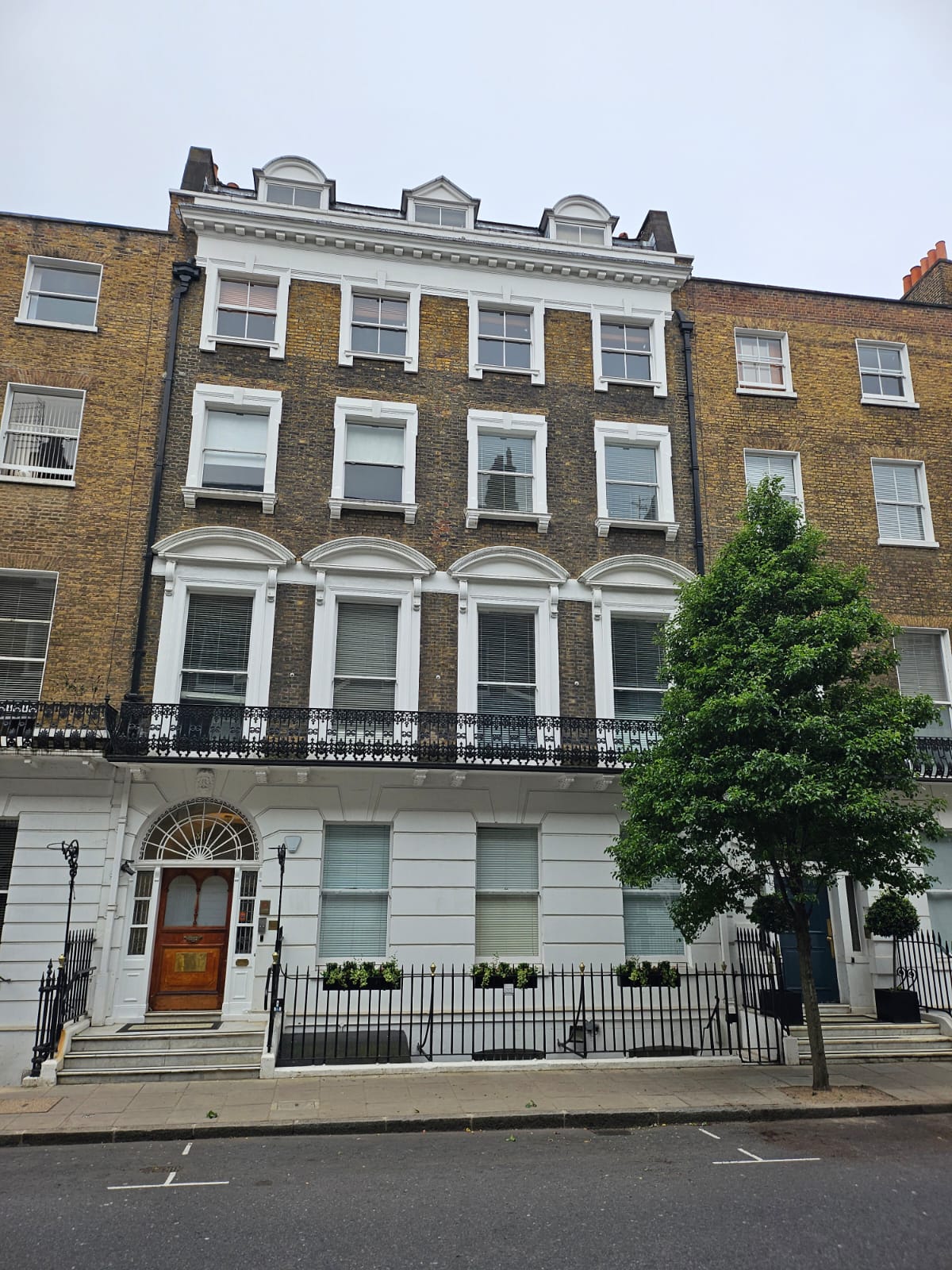 81 Harley Street is typical London townhouses, with four stories and attics, rectangular Georgian windows and a plastered ground floor and brick above. There is a tree growing on the street, on the right hand side as you look at the house.
